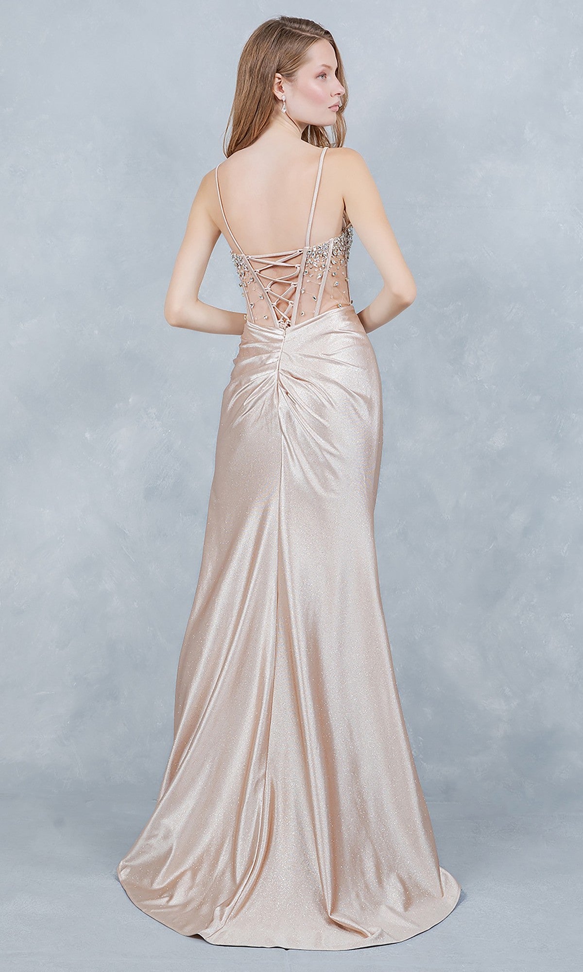 A woman wears the Cinderella Couture 8562J long prom dress, featuring a shimmery champagne hue, jeweled corset bodice, and lace-up back, standing before a light gray textured background.